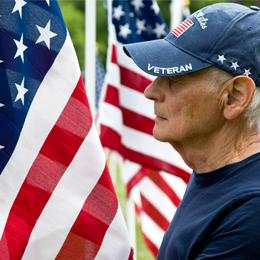 Older man wearing a U.S. veteran cap with American flags in the background